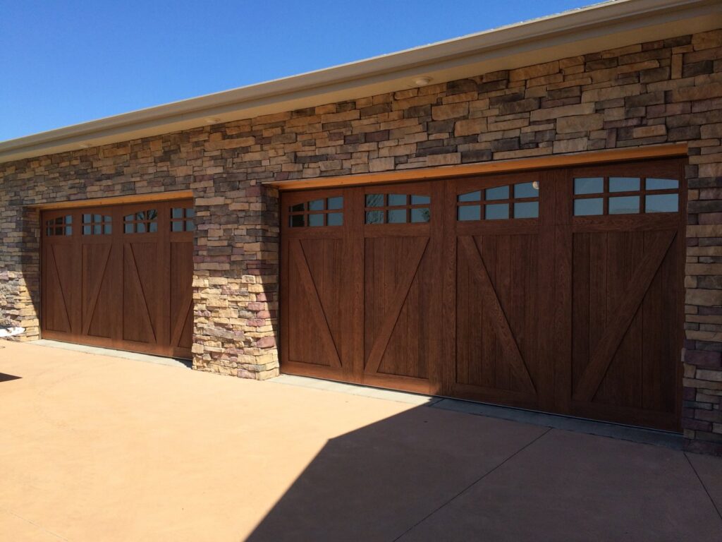 Two custom wood-look garage doors with decorative windows installed on a stone house by Alberts Custom Door Company in Wichita, KS.