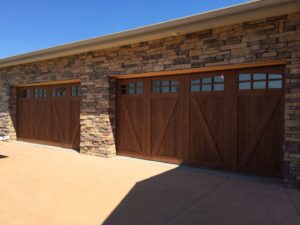 Two custom wood-look garage doors with decorative windows installed on a stone house by Alberts Custom Door Company in Wichita, KS.