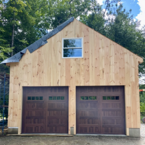 Two dark brown carriage-style garage doors installed on a light wood-sided building by Champion Overhead Door in Brattleboro, VT.