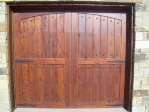 Two finished wooden carriage-style garage doors with decorative hardware installed on a stone house by Alberts Custom Door Company in Wichita, KS.
