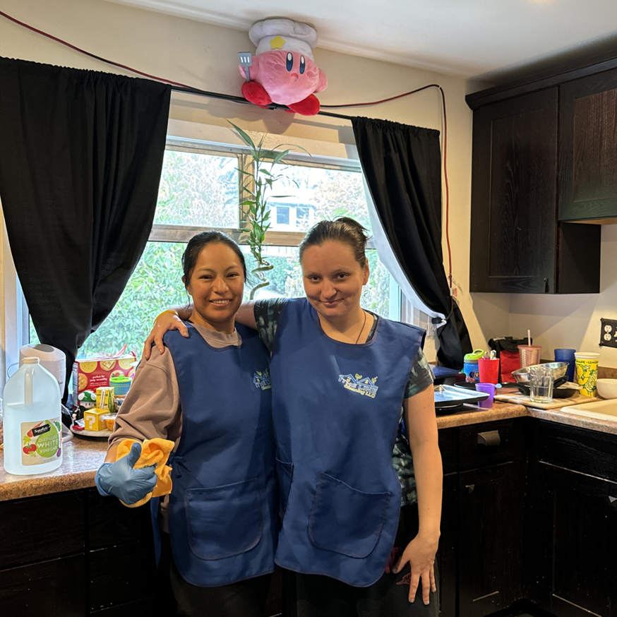 Two professional house cleaners from The Line Cleaning LLC in Renton, WA, smiling in a kitchen with cleaning supplies.