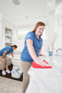 Two Maid Brigade cleaners in uniform wiping a bathroom sink and cleaning a bathtub in a home in Houston, TX.