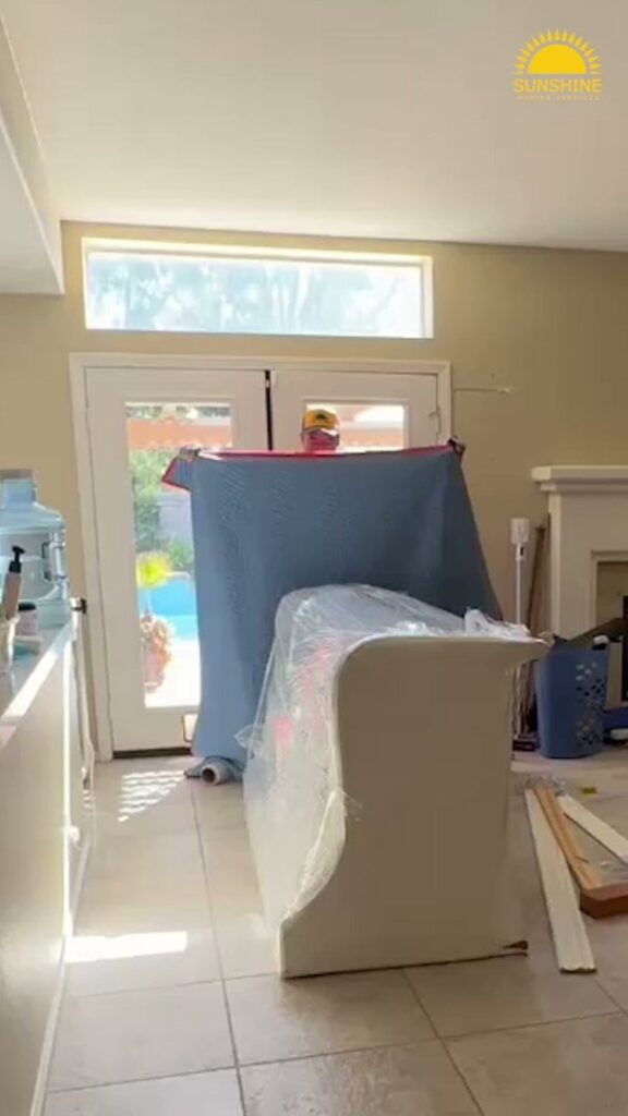 Two professional movers from Sunshine Moving Services LLC carefully carrying a large, wrapped item through a doorway in San Diego, CA.