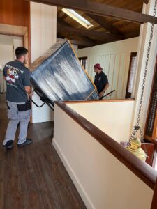 Two professional movers carefully carrying a large, wrapped item up an indoor staircase for All Way Moving in Washington, PA.