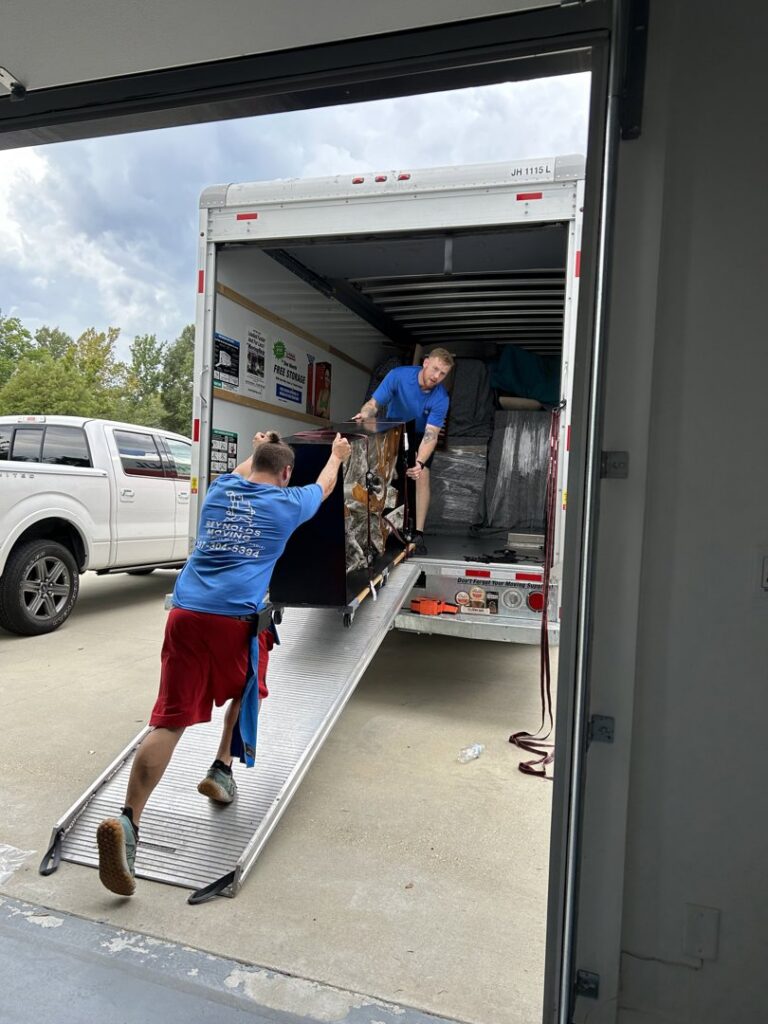 Two professional movers from Reynolds Moving in Baton Rouge, LA, carefully loading a large, wrapped item onto a moving truck via a ramp