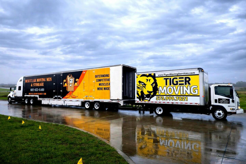 Two large moving trucks, one from Muscular Moving Men & Storage, parked on a wet road in Phoenix, AZ.