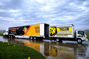 Two large moving trucks, one from Muscular Moving Men & Storage, parked on a wet road in Phoenix, AZ.