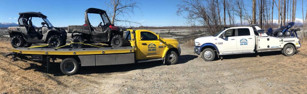 Two Help Line Towing trucks, one flatbed transporting two ATVs, parked in a rural area in Wasilla, AK.