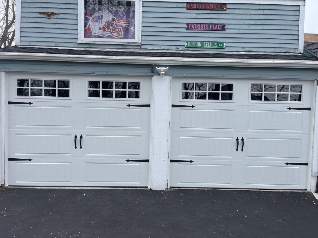 Two white carriage-style garage doors with decorative black hardware on a blue house, installed by Steve Shumsky Unlimited Overhead Doors LLC in Hudson, NH