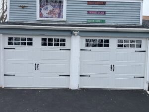 Two white carriage-style garage doors with decorative black hardware on a blue house, installed by Steve Shumsky Unlimited Overhead Doors LLC in Hudson, NH