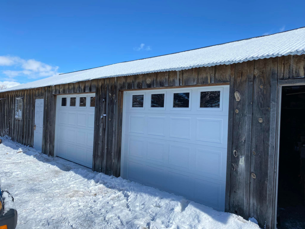 Two newly installed white garage doors with windows on a rustic wooden building by MBP Garage Doors in Firth, ID