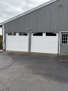 Two white panel garage doors with windows installed on a grey-sided building by The Garage Door Guys, LLC in New Haven, CT.