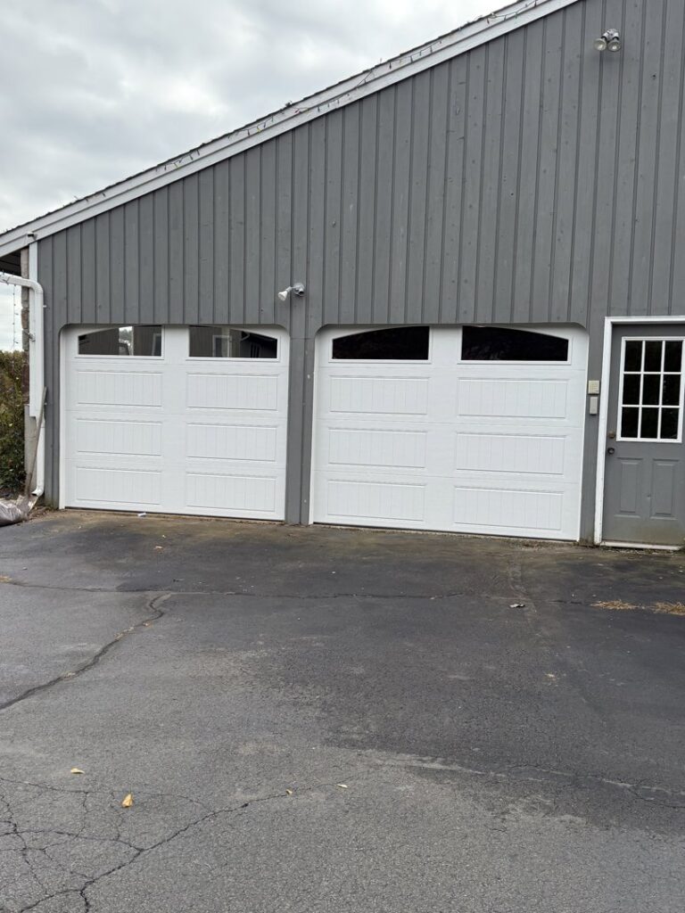 Two white panel garage doors with windows installed on a grey-sided building by The Garage Door Guys, LLC in New Haven, CT.