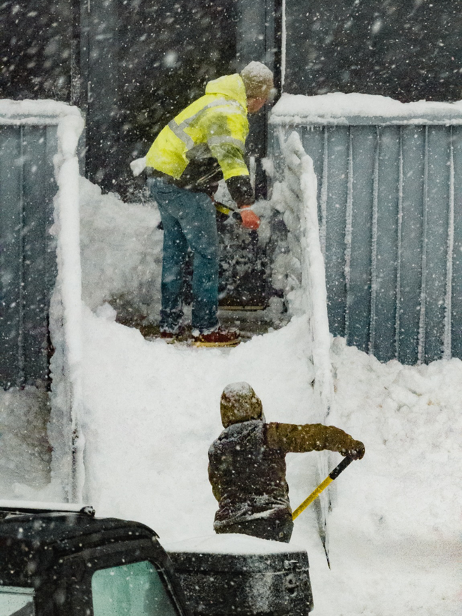 Two workers manually shoveling snow from different levels for Elcor Construction in Rochester, MN.
