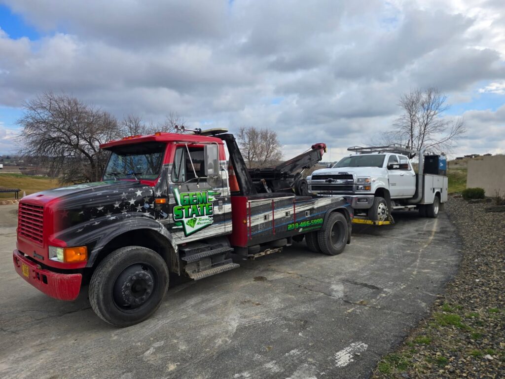 A white utility truck being towed by a wrecker from Gem State Towing and Recovery in Caldwell, ID.