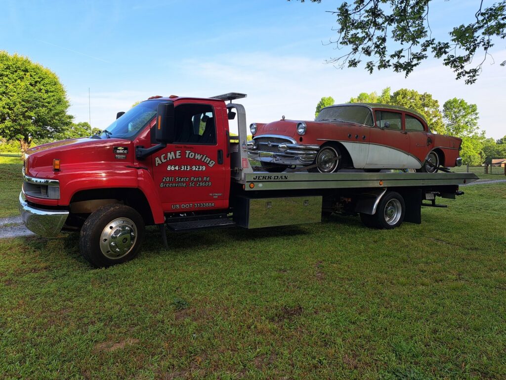 A vintage red and white car being transported on a flatbed tow truck by Acme Towing Services in Greenville, SC.