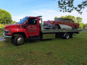 A vintage red and white car being transported on a flatbed tow truck by Acme Towing Services in Greenville, SC.