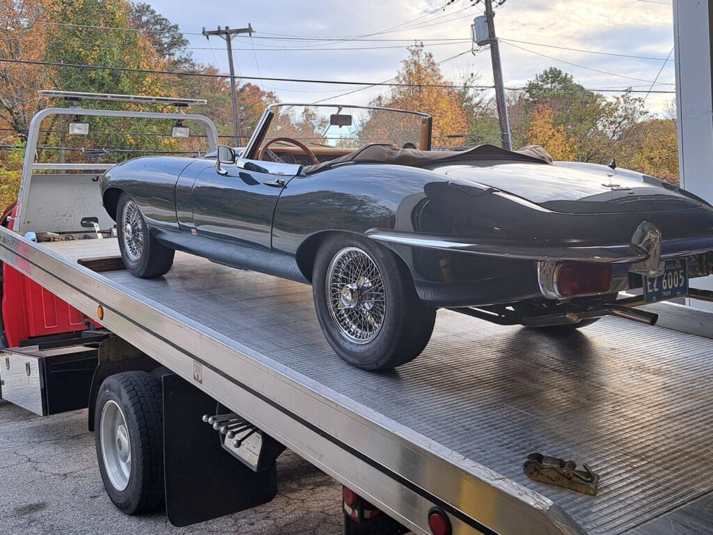 A vintage dark green Jaguar convertible being transported on a flatbed tow truck by Acme Towing Services in Greenville, SC.