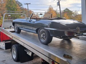 A vintage dark green Jaguar convertible being transported on a flatbed tow truck by Acme Towing Services in Greenville, SC.