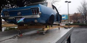 A vintage blue Mustang, without wheels and on dollies, being transported on a flatbed tow truck by Acme Towing Services in Greenville, SC.