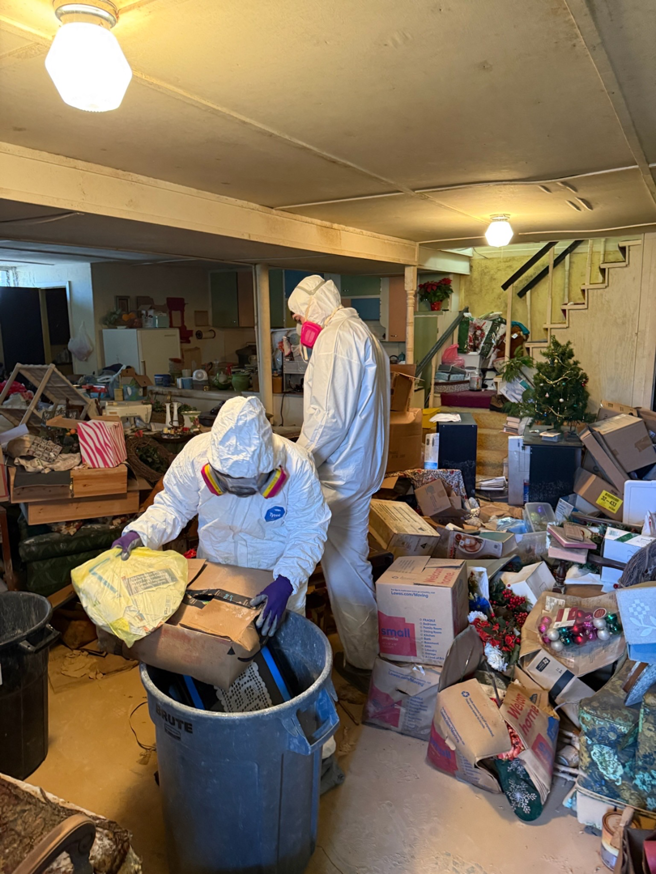 Two workers in hazmat suits cleaning a cluttered basement after water damage, performing remediation for Elam Enterprises in Jonesboro, AR.
