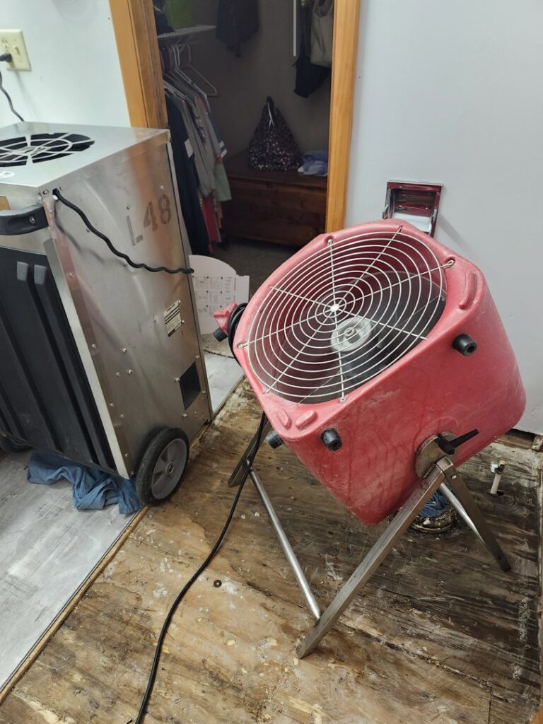 Water damage restoration equipment, including an air mover and dehumidifier, drying out a room with exposed subflooring by Magna-Dry in Lafayette, IN