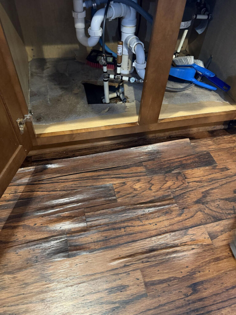 Water-damaged flooring under a sink cabinet, showing warping and discoloration, by Triangle Restoration in Raleigh, NC.