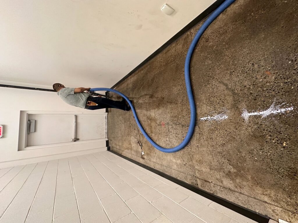 A specialist performing water extraction on a concrete floor using a large blue hose for The Recover Tech Restoration in Vancouver, WA.