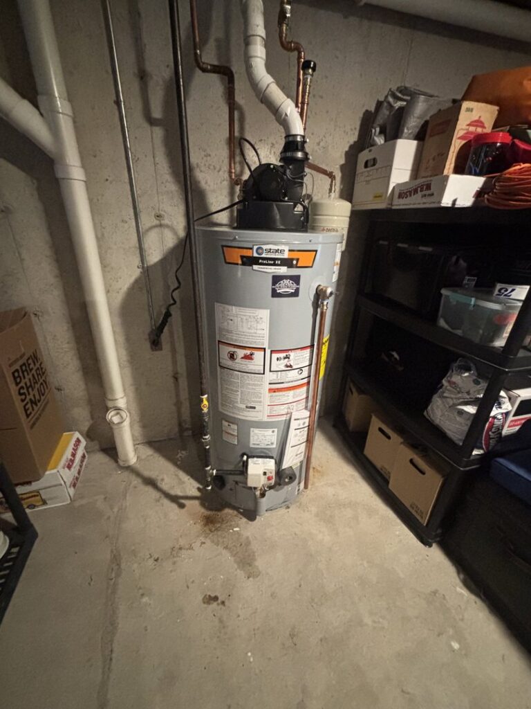 A water heater with a visible puddle and stains on the floor, indicating a leak and water damage, by White Birch Plumbing in Derry, NH.