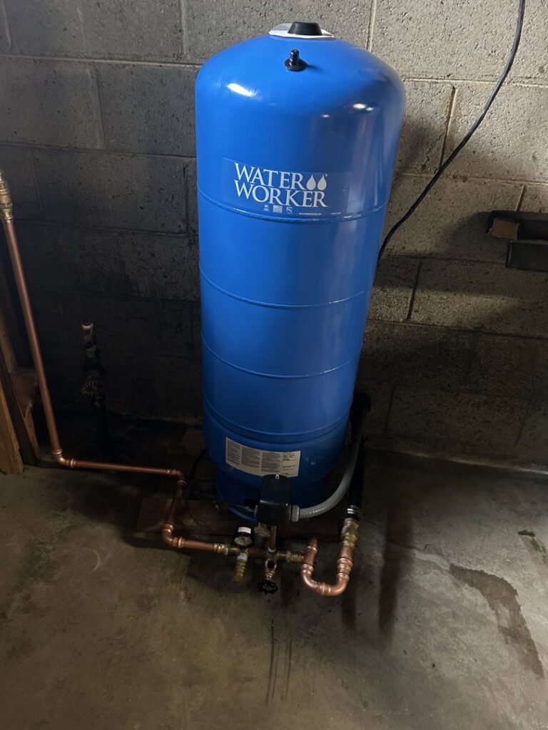 A 'Water Worker' tank and plumbing in a basement with damp floor, indicating water damage service by Burt Mechanical in Flint, MI.