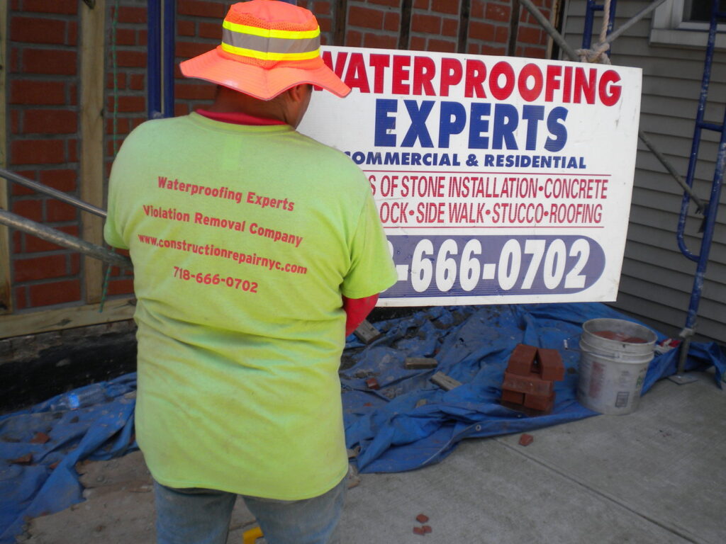 A worker wearing a 'Waterproofing Experts' shirt on a job site for Construction Repair NYC in Jamaica, NY