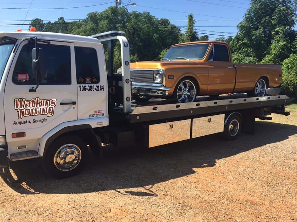 A Watkins Towing flatbed truck transporting an orange classic pickup truck in Augusta, GA.