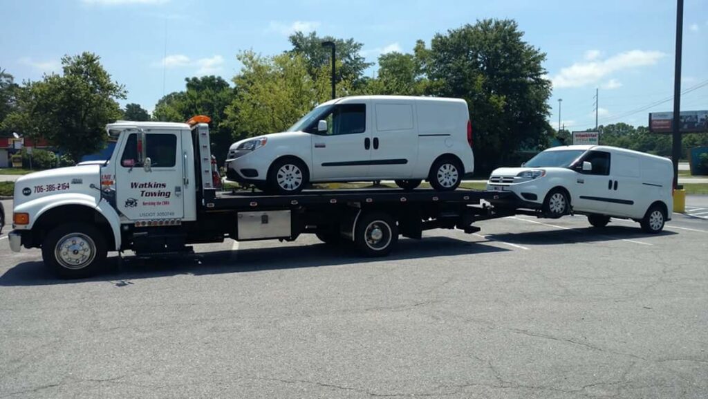 A Watkins Towing truck transporting two white vans, one on the flatbed and one towed behind, in Augusta, GA.