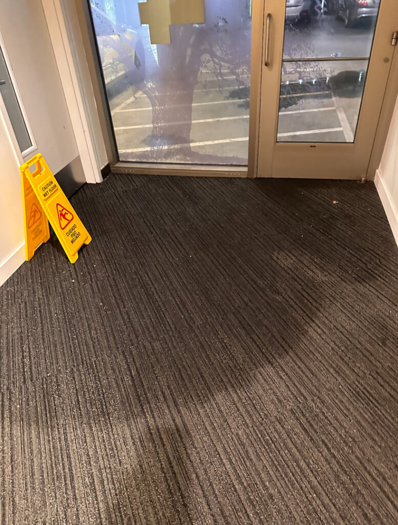 An entrance area with wet carpet and 'Caution Wet Floor' signs, indicating a water issue, for The Recover Tech Restoration in Vancouver, WA.