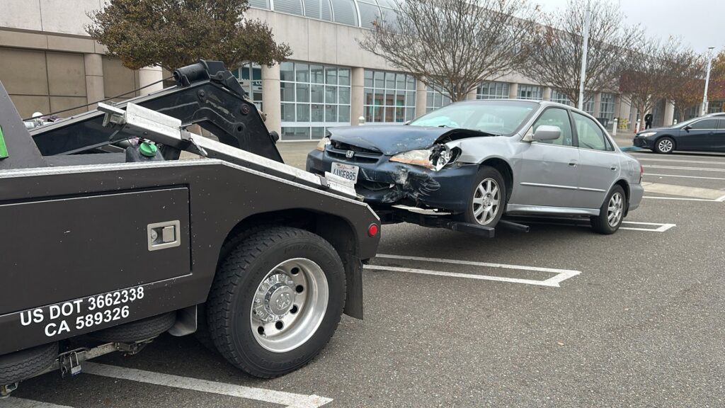 A damaged silver car being towed by a wheel lift tow truck by R towing Services in Sacramento, CA.