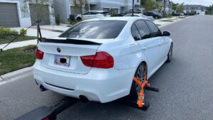 A white BMW sedan being towed by a TP Towing wheel lift truck on a residential street in Tampa, FL.
