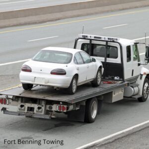 A white car being transported on a flatbed tow truck on a highway by Fort Benning Towing in Columbus, GA