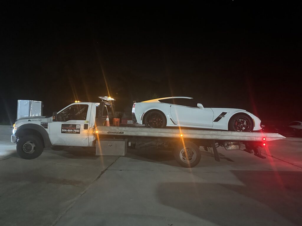TJ Towing LLC flatbed truck towing a white Corvette sports car at night in New Orleans, LA.