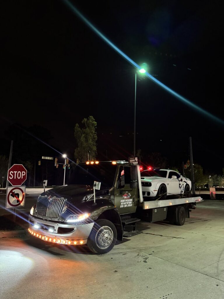 A white Dodge Challenger loaded onto a flatbed tow truck from Bullcity Towing & Services LLC at night in Durham, NC.