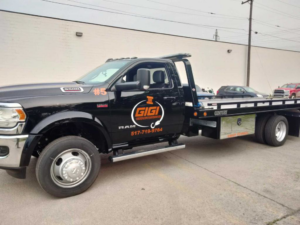 A white flatbed tow truck from Gigi Transportation carrying a dark grey pickup truck on its bed in Grand Rapids, MI.