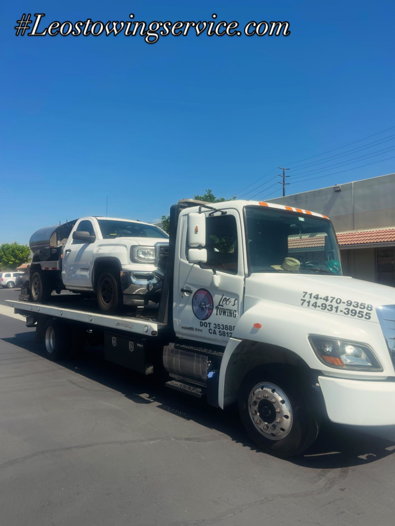 A white flatbed tow truck from Leos Towing Service carrying a white pickup truck in Anaheim, CA.