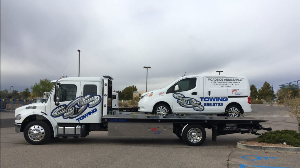 A white flatbed tow truck from SOS Towing carrying a white service van in Albuquerque, NM.