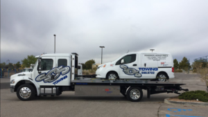 A white flatbed tow truck from SOS Towing carrying a white service van in Albuquerque, NM.