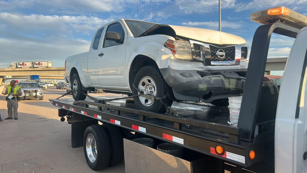 A white flatbed tow truck from Power Towing & Transport towing a damaged white pickup truck on a highway in Phoenix, AZ.