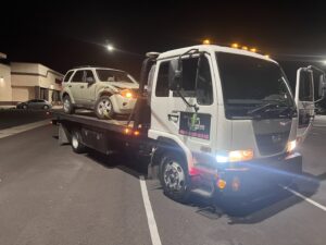 A white flatbed tow truck from Power Towing & Transport towing a damaged beige SUV at night in Phoenix, AZ.