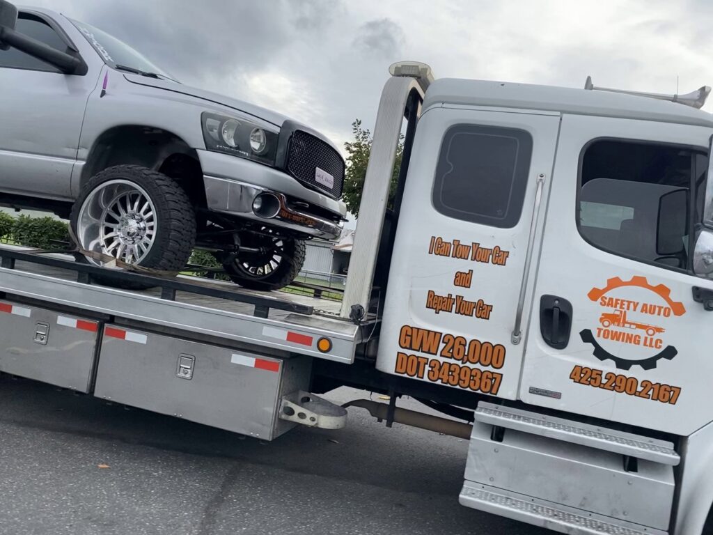A white flatbed tow truck from Safety AUTO Towing LLC in Seattle, WA, transporting a silver pickup truck.