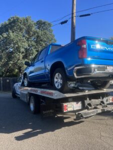 A white flatbed tow truck from Kennedy Towing & Transport in Anderson, CA, transporting a blue Chevrolet Silverado pickup truck.