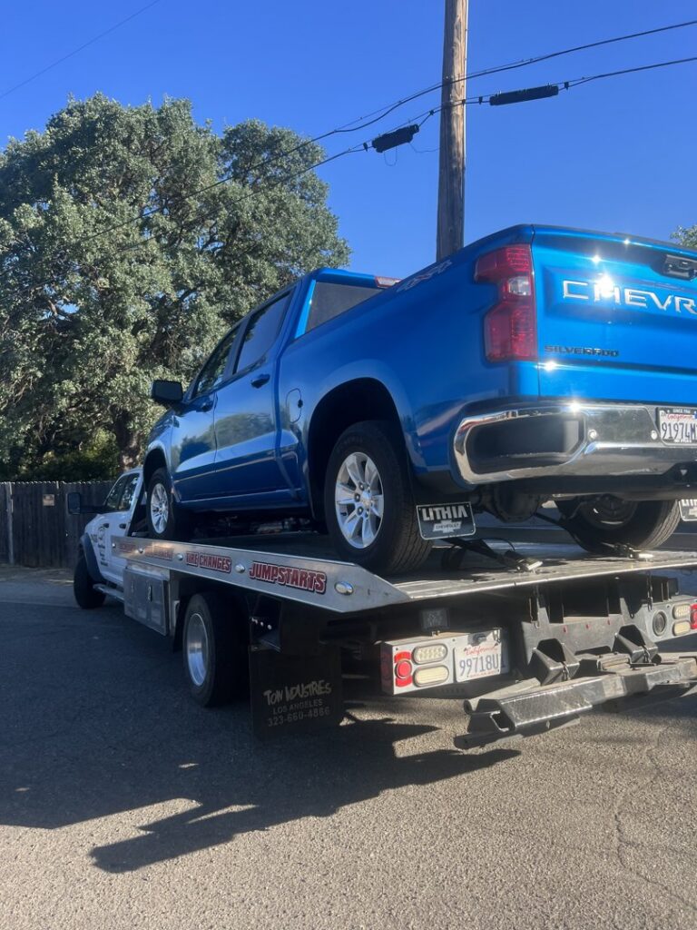 A white flatbed tow truck from Kennedy Towing & Transport in Anderson, CA, transporting a blue Chevrolet Silverado pickup truck.