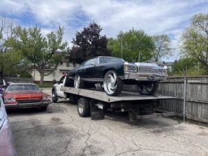 A white flatbed tow truck transporting a dark classic car for Boyz Automotive & Towing in Toledo, OH.