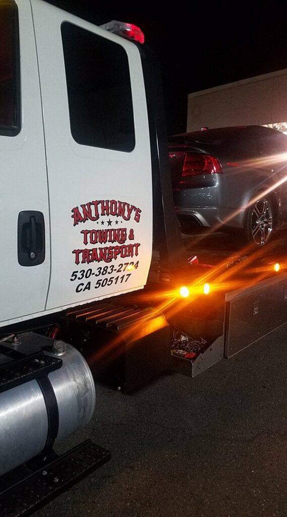 A white flatbed tow truck from Anthony's Towing & Transportation transporting a dark car at night in Yuba City, CA.